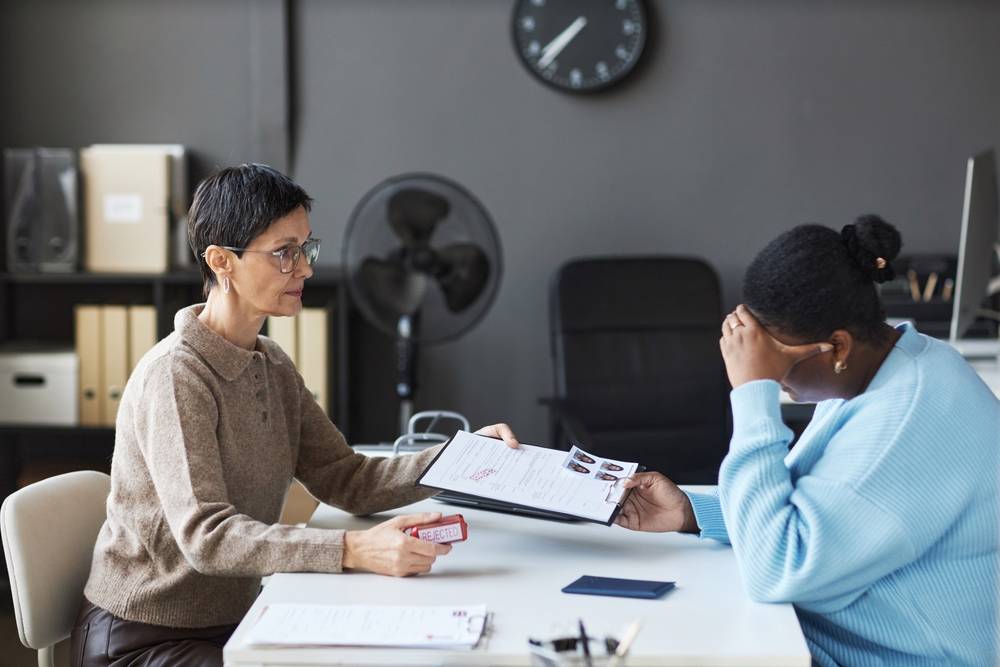 UK Visitor Visa Application: Side view of mature female manager passing rejected visa application form to anxious black woman touching her head while feeling upset.