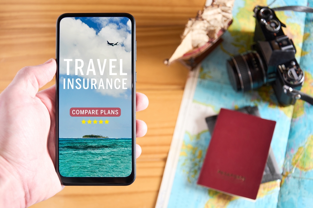 Travel Insurance for Canada: Top view of male hand holding mobile phone with travel insurance website on the screen. Travel items on table in the background with camera, passport and map